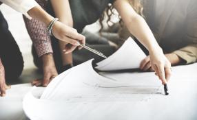 Image of people leaning over building plans on a table, one person is pointing to a document with a silver pen