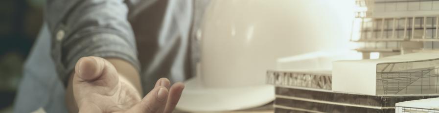 Image of a person pointing to a document and holding a white hard hat, leaning against a table