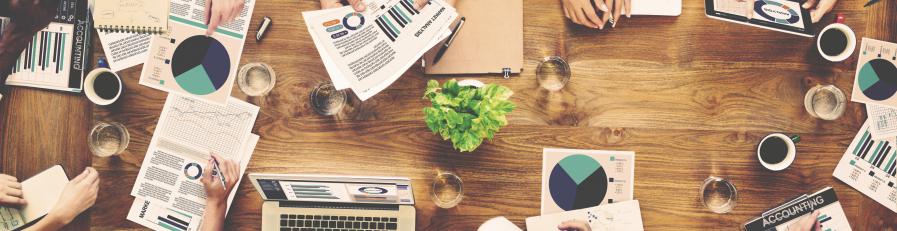 Image of a people sitting at a long table with paperwork, laptops, coffee and folders